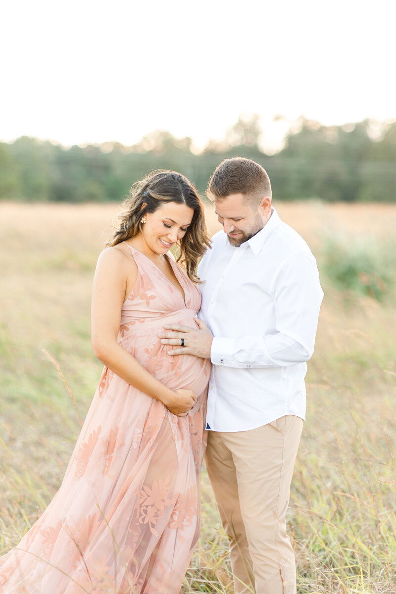 A DC Maternity Photographer of a pregnant couple in a tall grass field at sunset holding the pregnant belly and smiling