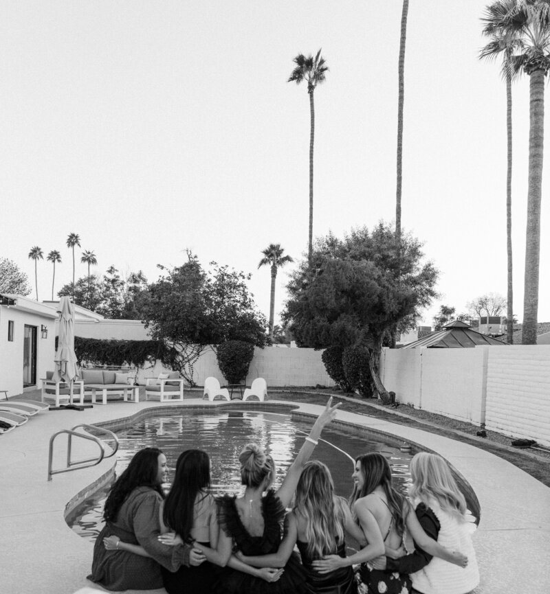 A group of six women sitting by a pool, facing away from the camera, with their arms around each other. One woman is raising her hand in a peace sign. The setting is a backyard with trees and patio furniture, creating a cozy and intimate atmosphere.