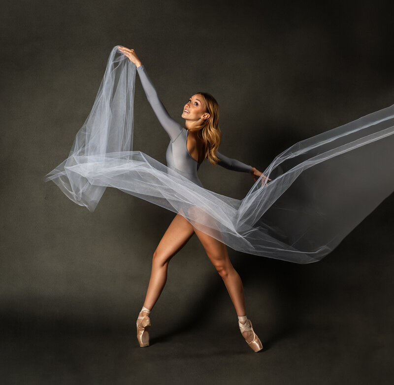 A ballerina in a gray leotard dances and twirls with a long piece of fabric  against a gray backdrop