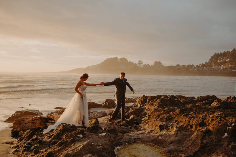 a woman in a gown has her hand outstretched to her groom on the rocky shores of Rockaway Beach on the Oregon Coast