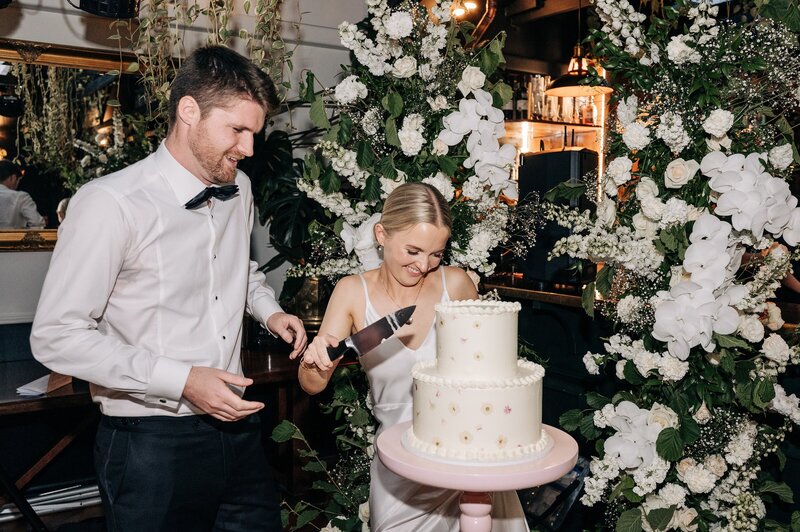 bride and groom cut fulltime tart wedding cake with white floral pillars at 5th street wedding reception restaurant in christchurch