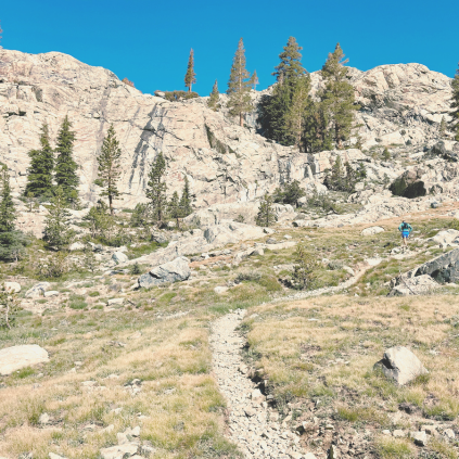 A man hikes a steep rocky trail in the John Muir Wilderness area of the Eastern Sierra Nevada. The sky is blue and rocky cliffs in the distance are light grey decorated with pine trees.