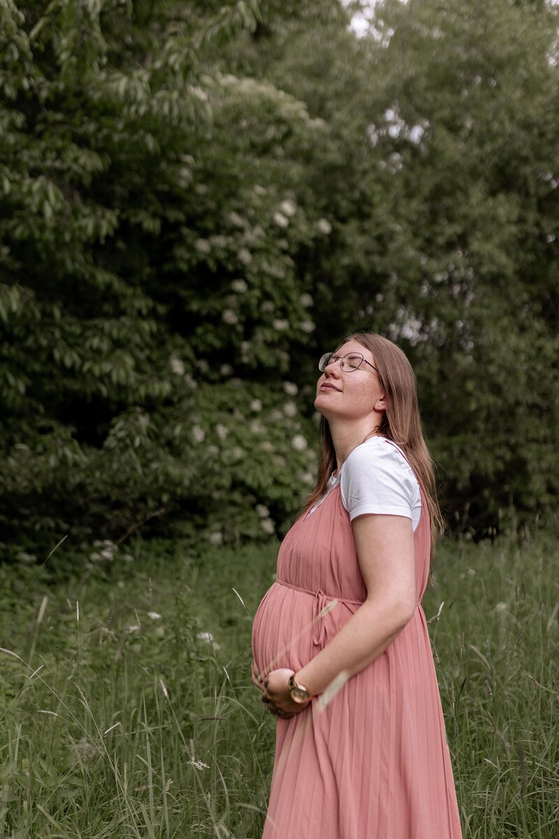 Frau steht auf einer Wiee vor Baeumen und haelt ihren Babybauch