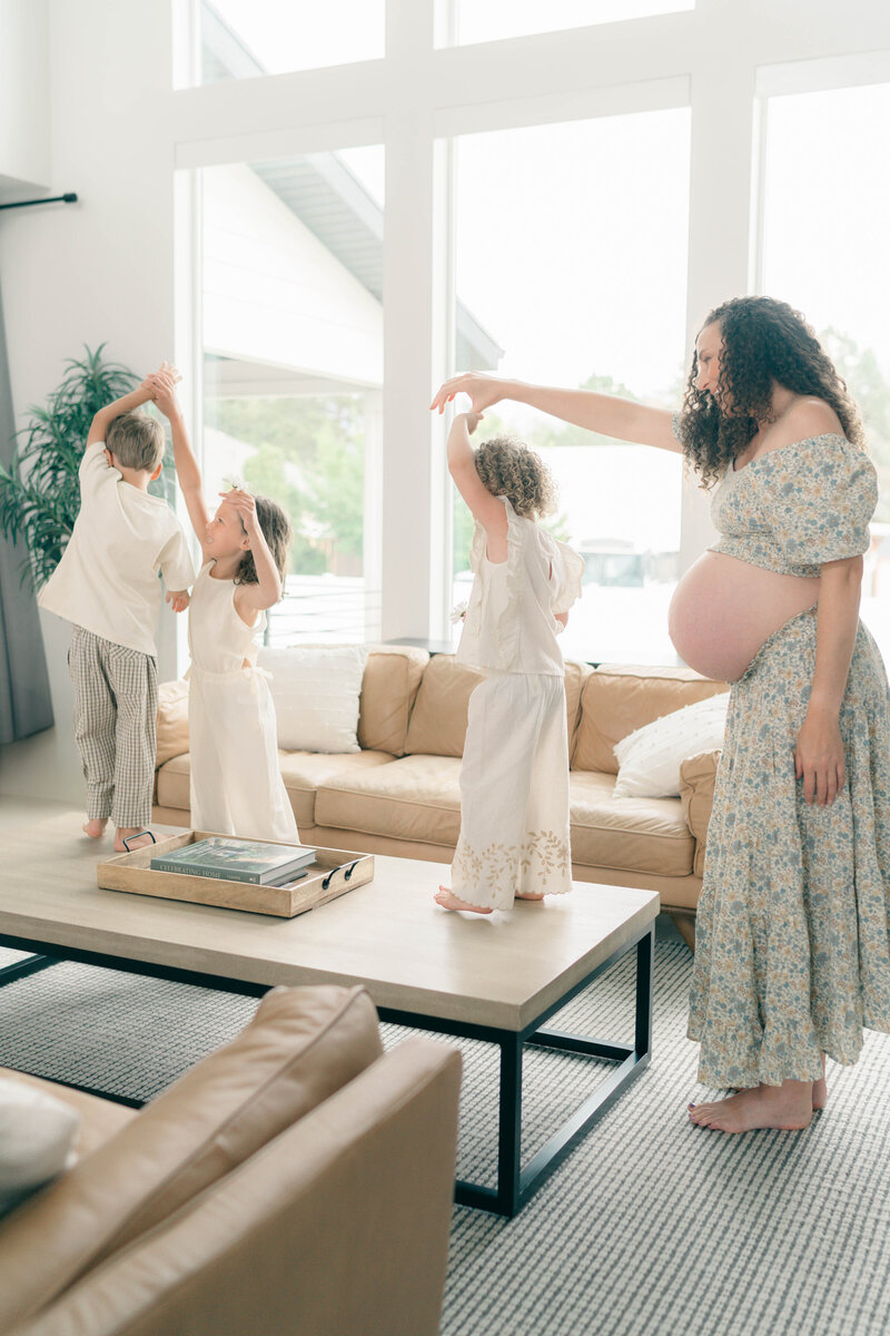 Riverton family photographer captures a cozy maternity session at home, with toddlers cuddling up to mom in a sun-drenched living room filled with natural light.