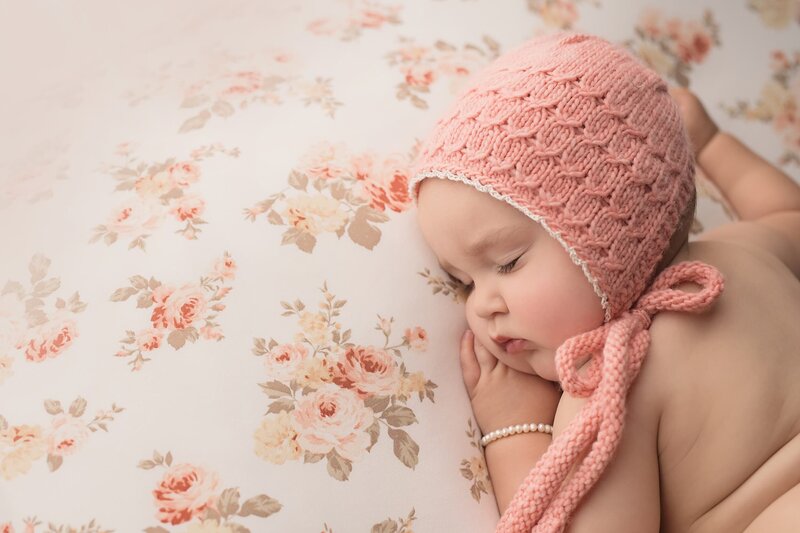 A newborn baby sleeps on a floral print bed in a pink knit bonnet and pearl bracelet