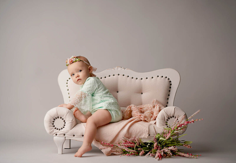 A baby in a mint green lace outfit sits on a small white tufted sofa, with a floral headband and pink flowers nearby, in a soft, serene setting.