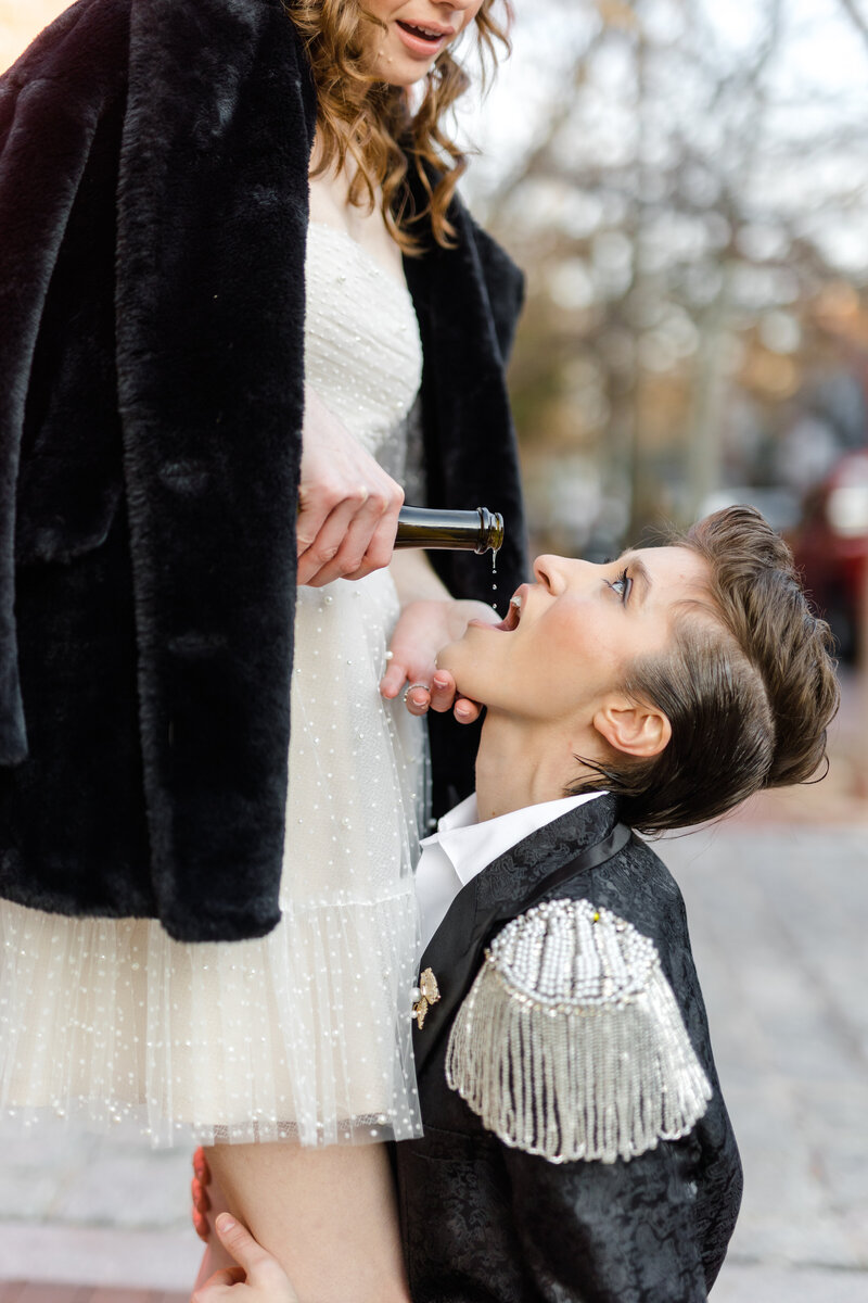 Queer bride in sheer tulle pearl mini dress pouring champagne into the mouth of her nonbinary partner who is kneeling in a black shimmer suit with pearl sequin tassel epaulets while they are staring up at them