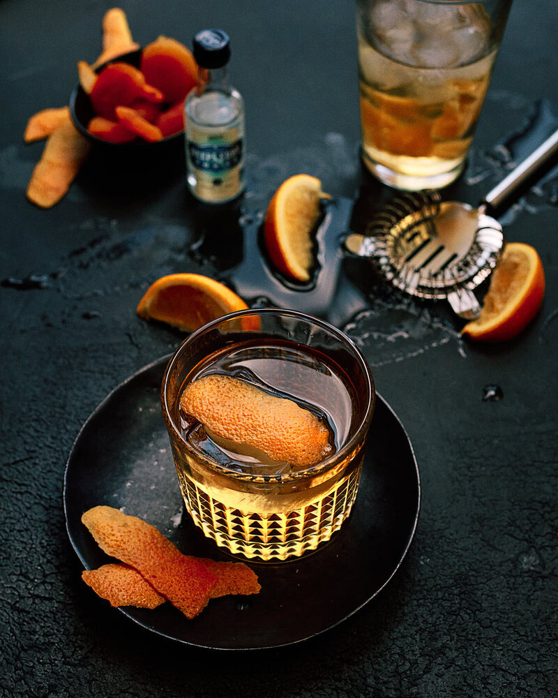 A rich and atmospheric image of a whiskey cocktail garnished with an orange peel, set against a dark background with scattered cocktail-making tools. Captured by Mica McCook, this photo highlights the craftsmanship and flavor of a well-made cocktail.