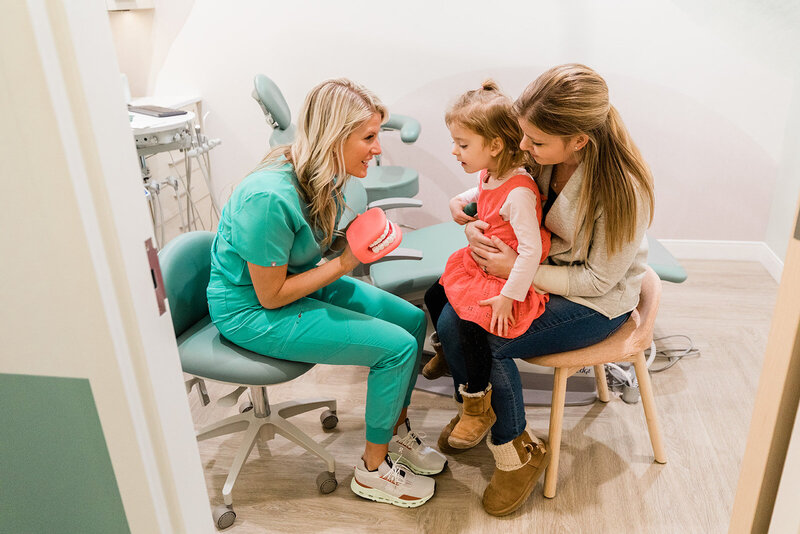 The dental hygienist for Dr. Michael Rabinowitz, DDS, shows a young patient a puppet of what her mouth and teeth look like. The pediatric dental patient looks with interest at the faux mouth, while her mom holds her on her lap. The pediatric dental exam area and tools can be seen in the background.