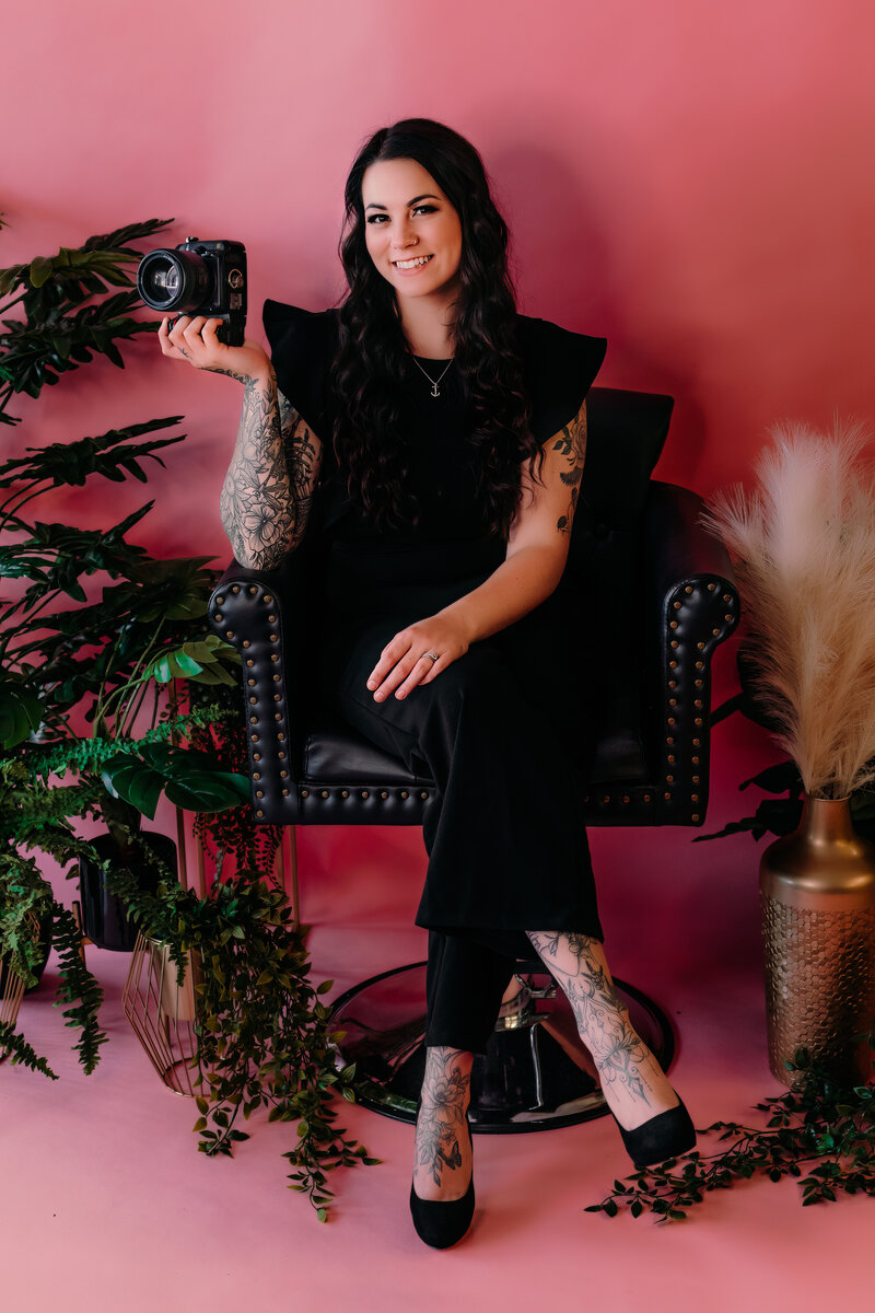 Portrait of Kendri, a photographer, sitting in a stylish chair against a vibrant pink background, holding a camera in her right hand. She is dressed in black, with plants and decorative elements surrounding her, conveying a warm and professional vibe. The text above reads, ‘Hi, I’m Kendri!