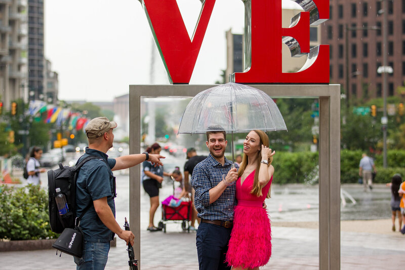 Engaged couple posing romantically in the rain in front of the iconic Love Statue during an engagement session, with Travis Stitt providing guidance to create a beautifully cinematic and heartfelt moment.
