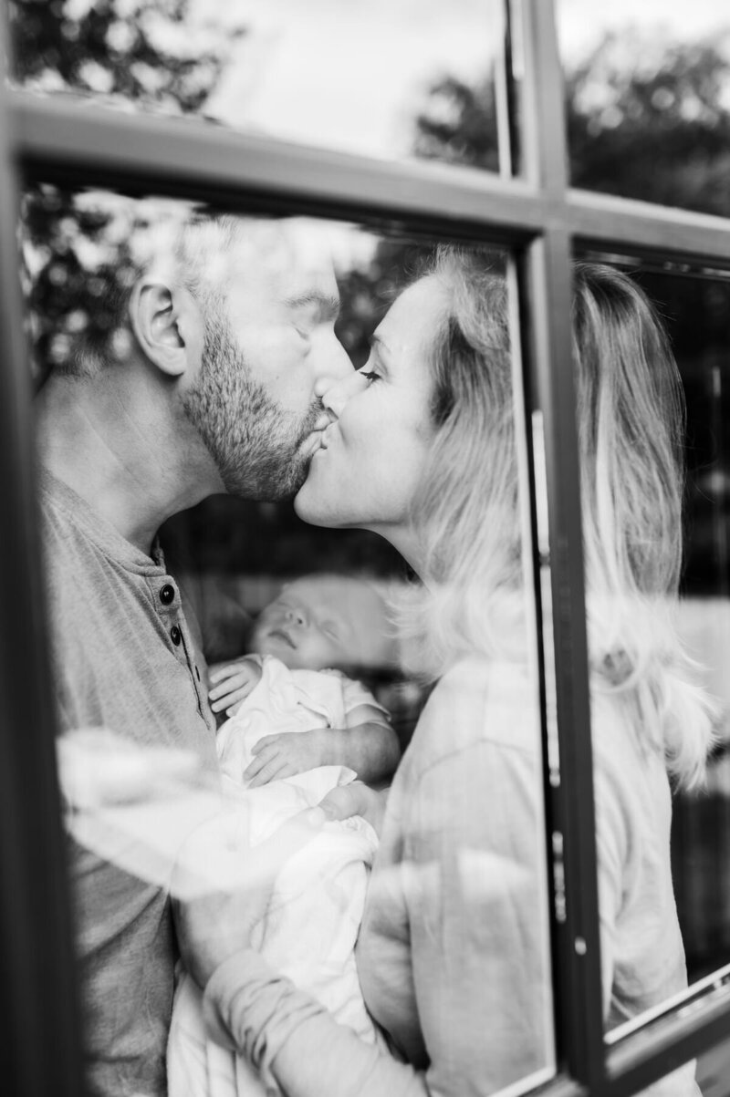 black and white photo taken through window of mom and dad holding baby and kissing