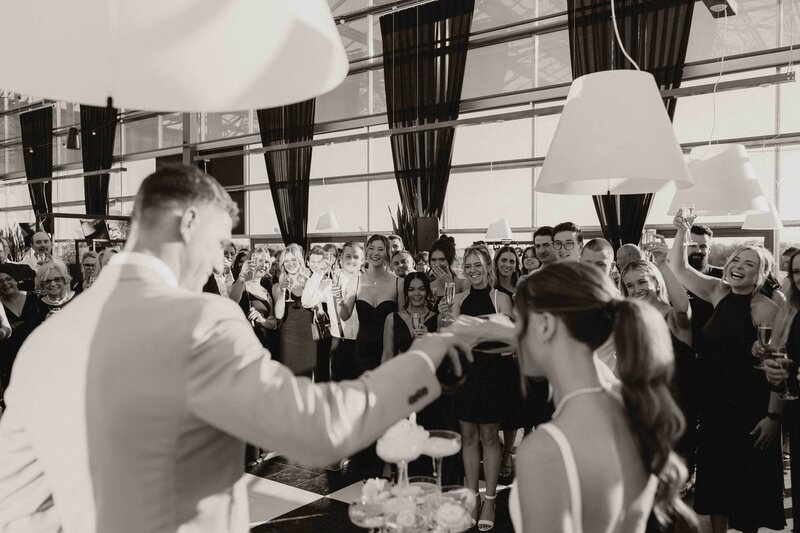 Black and white photo shot from behind, showing bride and groom pouring champagne onto a tower in front of their standing guests.