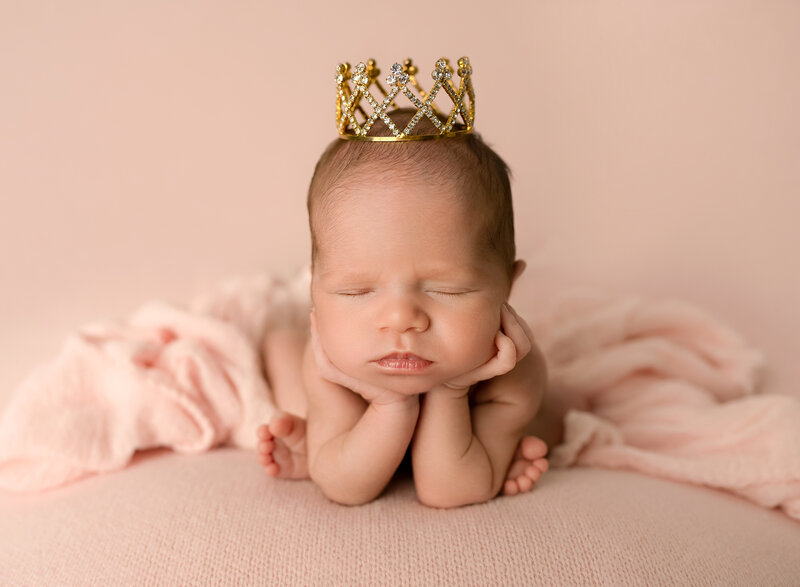 newborn girl wearing gold crown in froggy pose on pink blanket