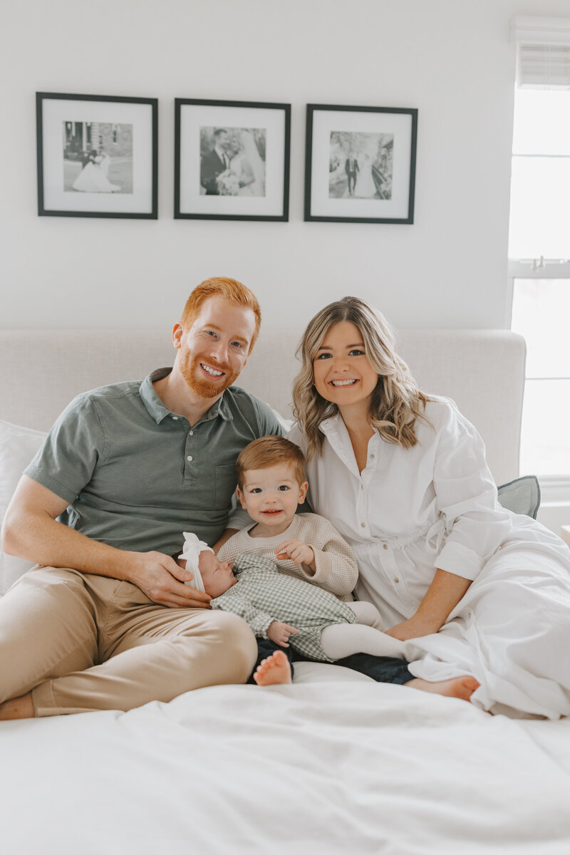 Family with son and newborn daughter smiling looking at camera while sitting on bed