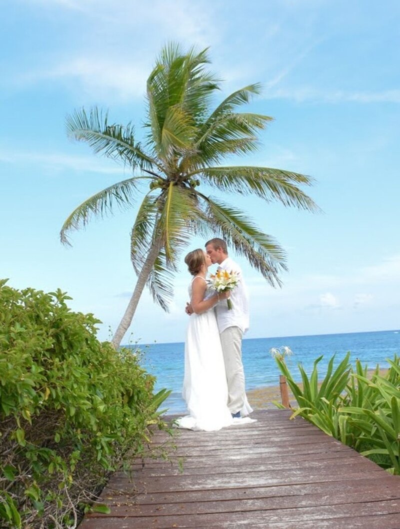 Bride and groom embracing on a tropical boardwalk overlooking the ocean, framed by lush greenery and a palm tree at their beach destination wedding