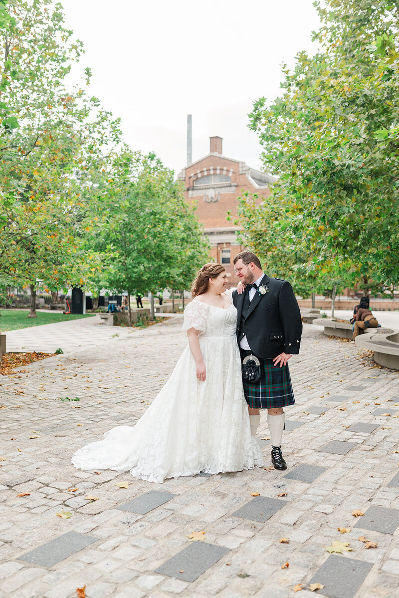 Jewish bride leaning on shoulder relaxed of Scottish groom outside on a white brick paved walkway in Navy Yard DC in front of brick industrial building 