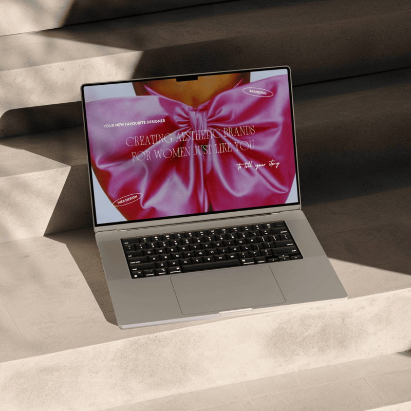 Close-up of a sleek laptop open on a set of concrete steps. The laptop screen displays a minimalist website design with the text "Your New Favorite Designer" and "Creating Aesthetic Brands for Women Just Like You.