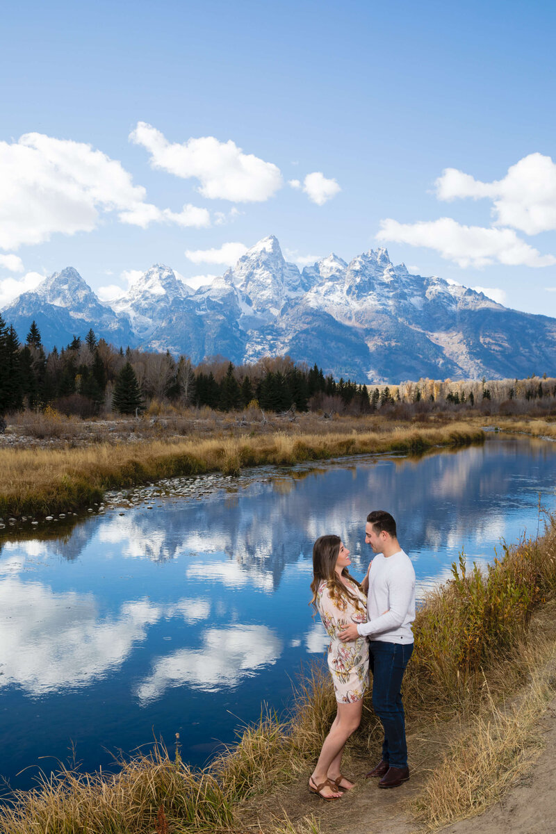 Couple shares a joyful embrace along the water at Schwabacher Landing, with the Grand Tetons reflecting in the still lake behind them.