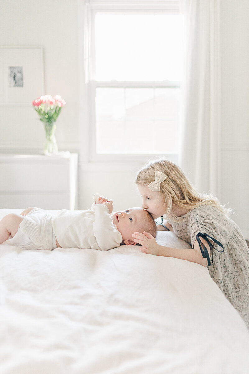 A toddler lovingly kisses her newborn sibling on a white bed during a peaceful in-home Kansas City newborn session by Meredith Maag Photography.