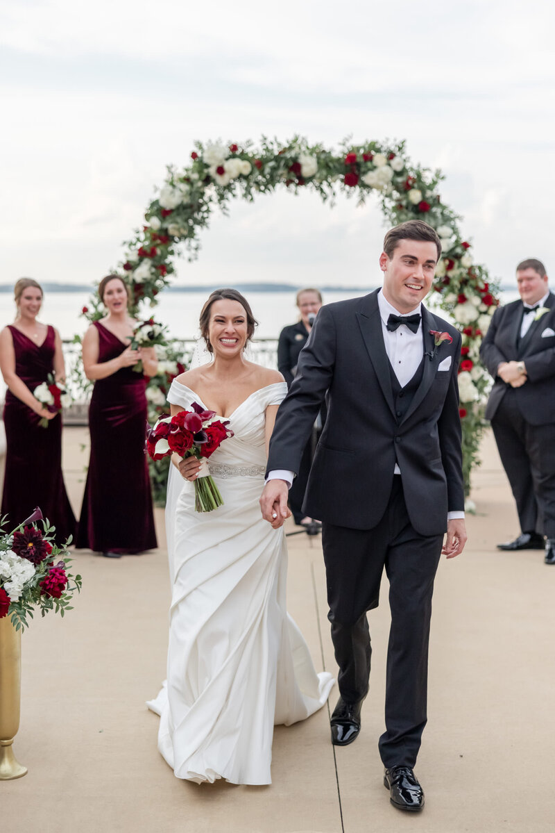 Bride and groom walk down aisle after getting married while holding a red floral bouquet