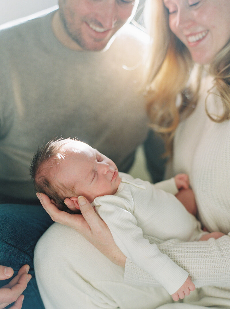 Newborn girl captured by Seattle Film Photographer backlit by sun in her parents arms at home in Seattle