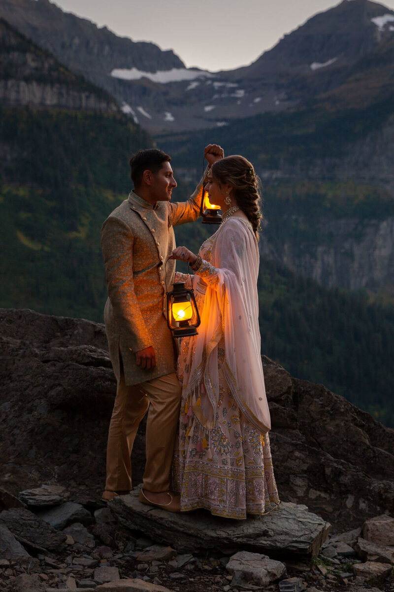 Couple in traditional South Asian wedding attire holding lanterns at dusk on a mountain ledge, gazing at each other as the warm glow illuminates their faces.