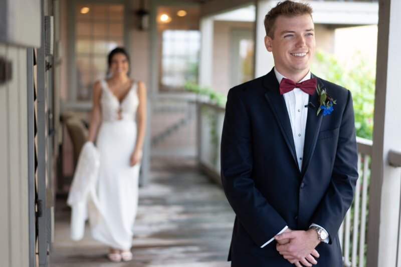 Groom in navy tuxedo with burgundy bow tie and blue boutonniere smiling during first look moment, with bride in white dress visible in soft focus background at the White Room wedding venue in historic downtown St. Augustine
