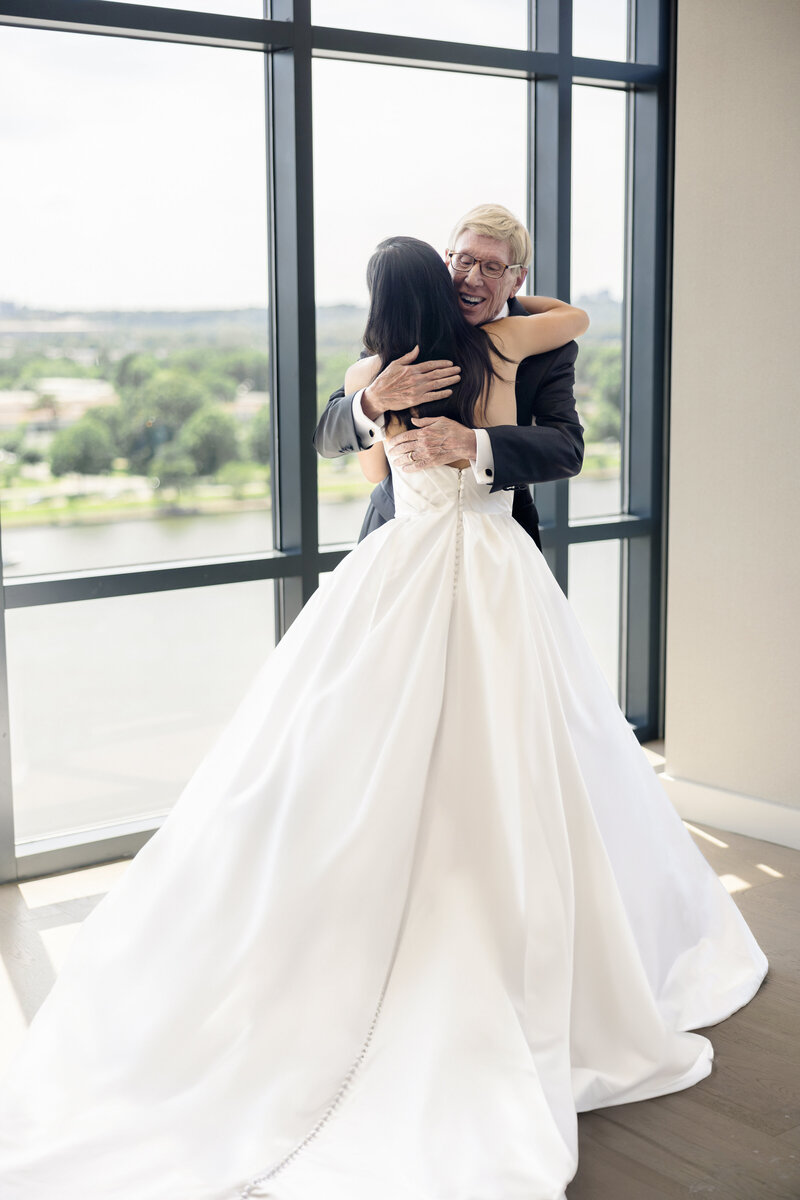 A bride in a white gown hugs an older man in a suit, standing by a window overlooking a scenic view with trees and a river. The room is filled with natural light, creating a warm and joyful atmosphere.