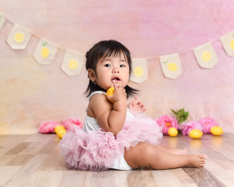A baby girl sitting on a wooden floor in a pink tutu, holding a yellow lemon toy with a lemon-themed banner and flowers in the background.