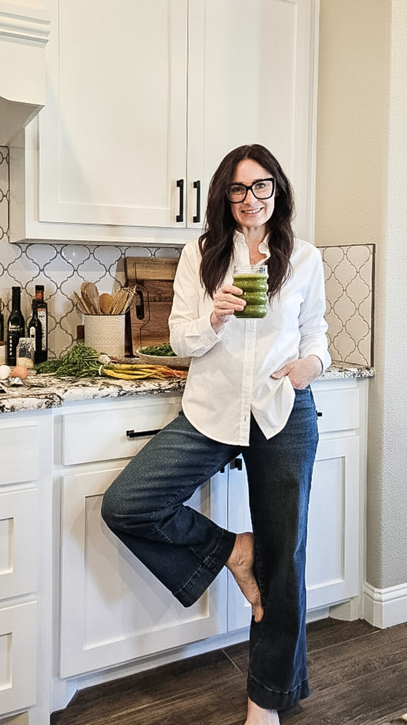 Woman with brown hair in a white button down shirt in a kitchen with white cabinets holding a green juice to start the morning off right.