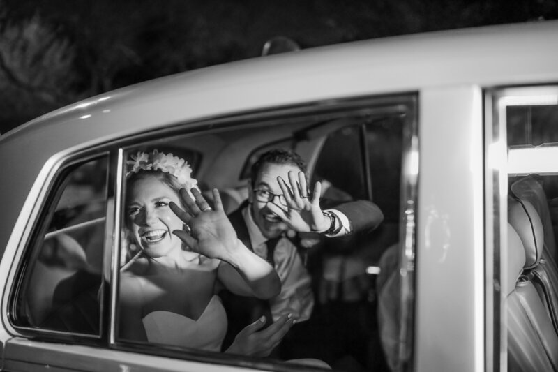 Black and white photo of bride and groom in their getaway car waving and surrounded by bubbles