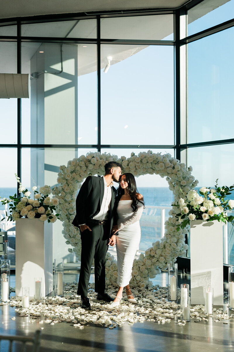 Couple posing in front of a white floral heart arch with rose petals and candles, overlooking the ocean, styled by Yours Truly, a wedding planner and designer in New Jersey.