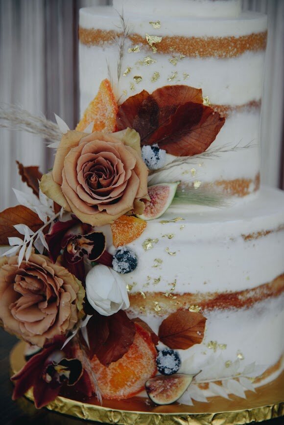 A close up shot of a tiered wedding cake. Naked style frosting with fall florals and gold leaf scattered.