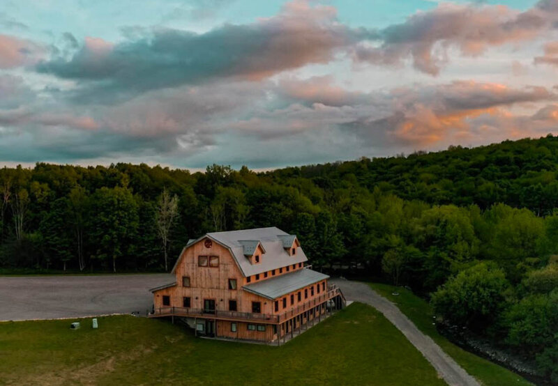 Aerial view of rustic barn venue surrounded by lush green forest under a moody sunset sky at Bear Ridge Destination.