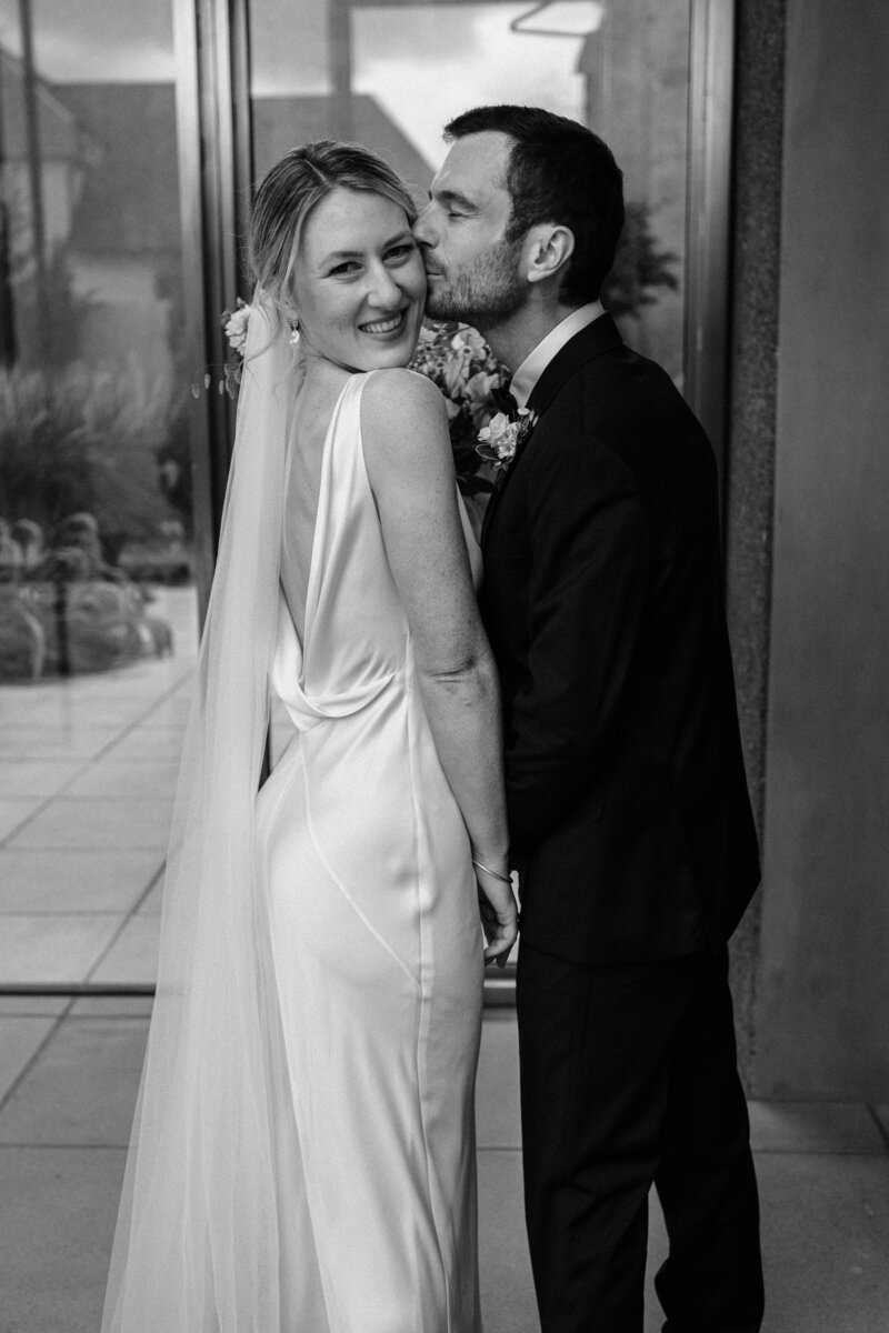 a black and white shot of bride with cheeky smile as groom kisses her in a candid moment in the christchurch cbd with an urban feel