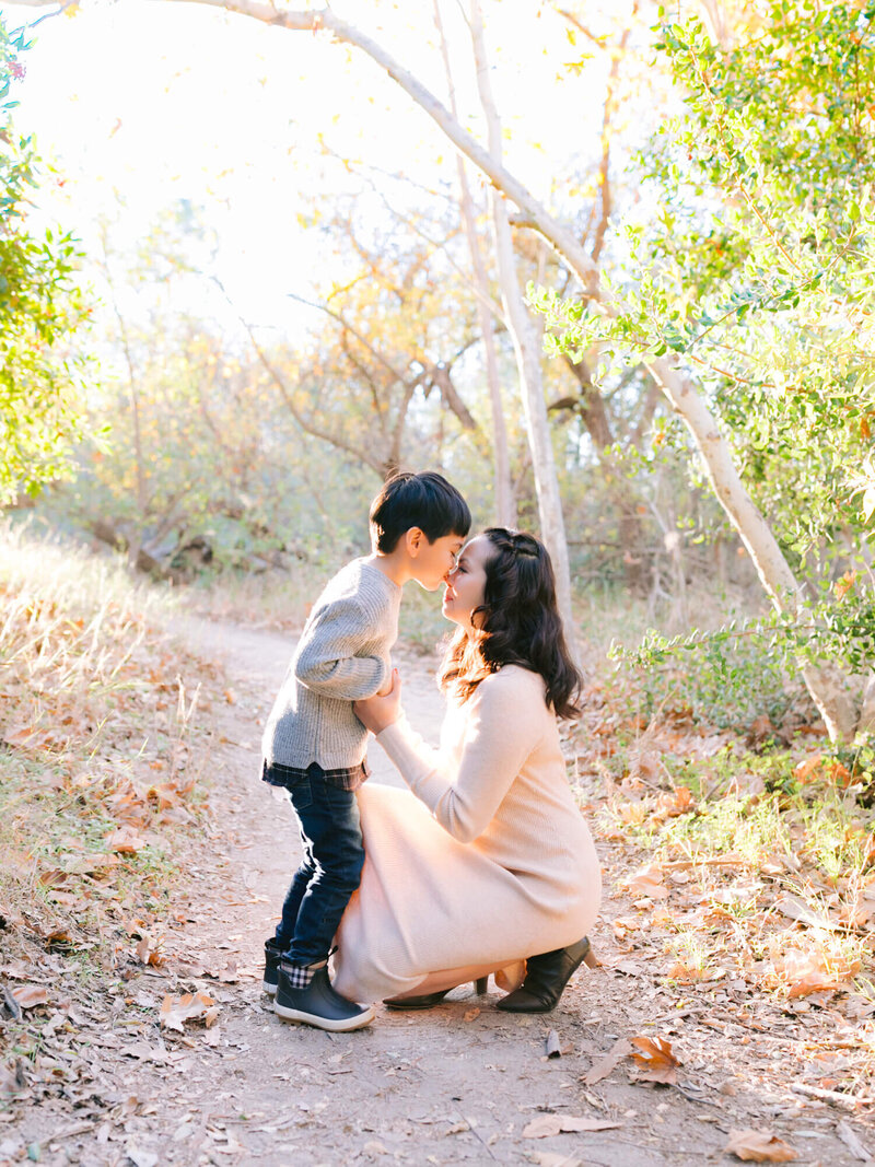 A mom kneels on a forest path, smiling as she touches noses with a young boy. Sunlight filters through the trees, creating a warm, tender atmosphere.