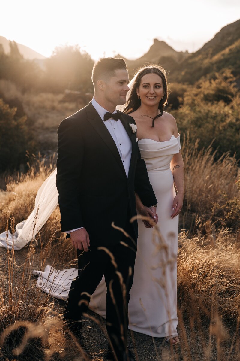 a bride smiles at her groom in the golden sunset in gold grass at the winehouse in queenstown