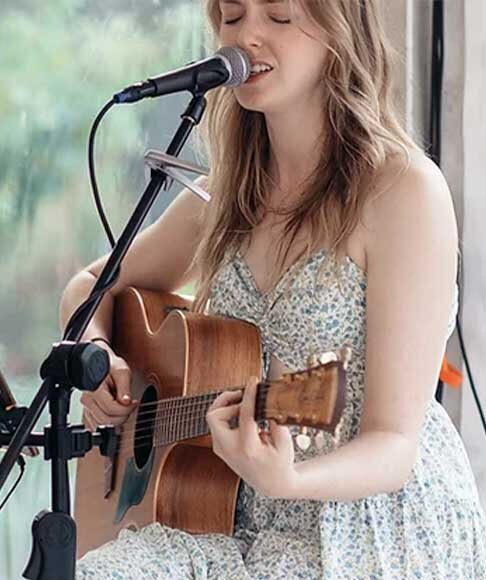 Female acoustic artist sings and plays guitar live on stage at an intimate daytime Coldplay event