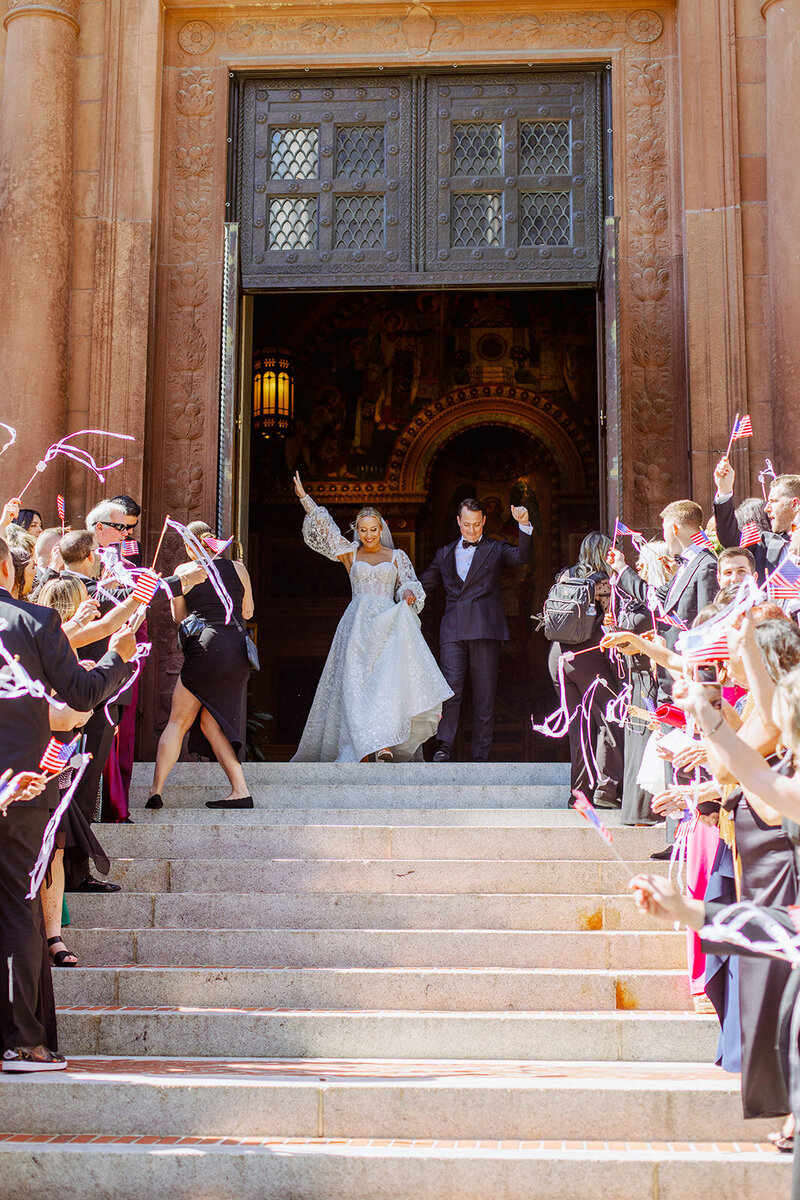 Bride and groom walk outside of St Matthews Church while guests cheer with flags