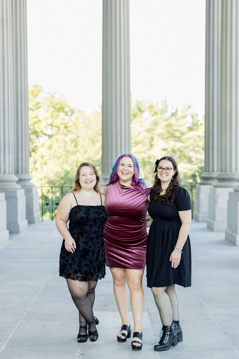 Jessica Hunt, owner of Jessica Hunt Photography, stands between two team members on a sunlit outdoor terrace lined with tall columns. Jessica is wearing a deep magenta velvet dress and black platform heels, with vibrant pink, purple, and blue hair. The team members on either side are dressed in black dresses, smiling brightly. The setting conveys a confident, stylish, and approachable creative team.