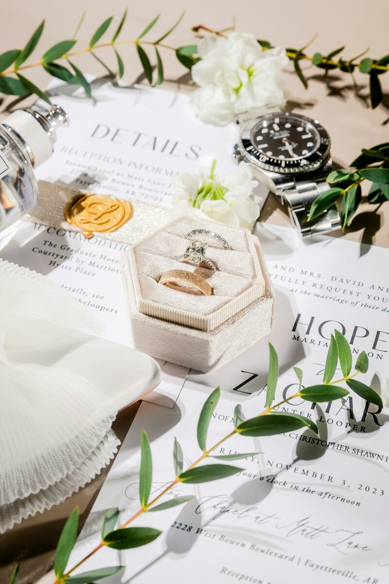 White Velvet ring box containing 3 rings, a men's watch, with greenery and white flowers, and wedding invitation suite in the background.