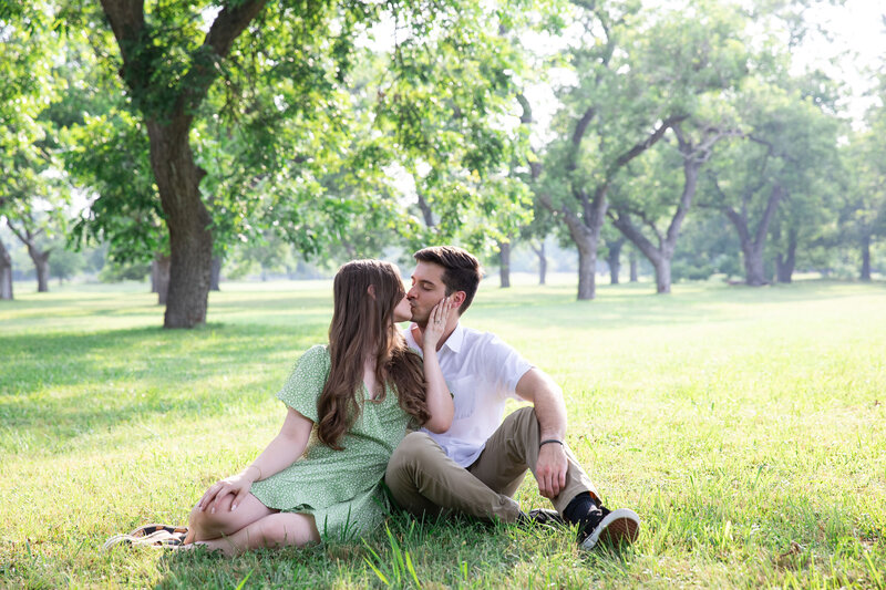A couple sits on the grass and kisses in a park, captured beautifully by an Austin wedding photographer.