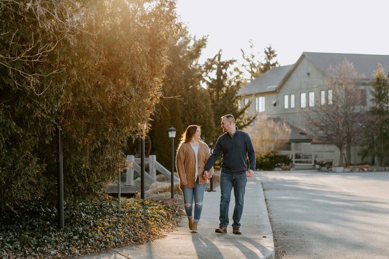 Engagement photoshoot at Bellemere Winery in London, Ontario. Man and woman are walking hand-in-hand along sidewalk in front of beautiful rustic building at golden hour.
