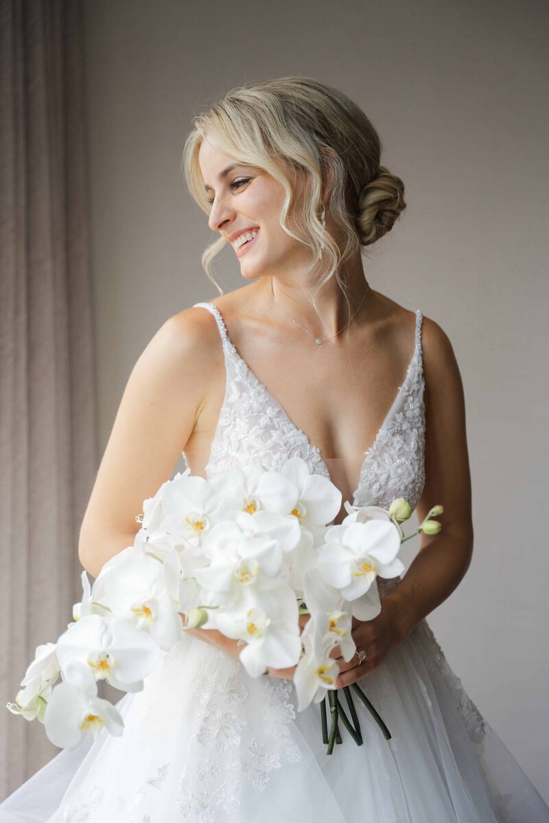 Bride in lace wedding gown holding a cascading white orchid bouquet, smiling softly in natural light.