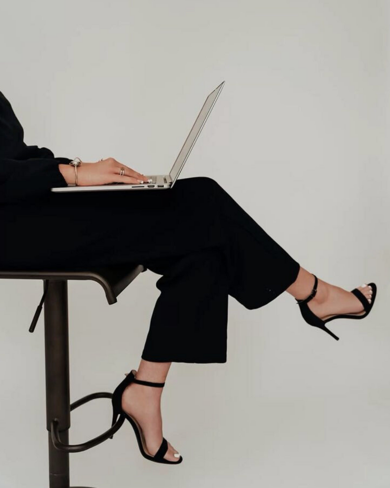 Editorial-style photo of Asmahan seated in black trousers and heels, working on a laptop with confident poise in a minimalist white studio