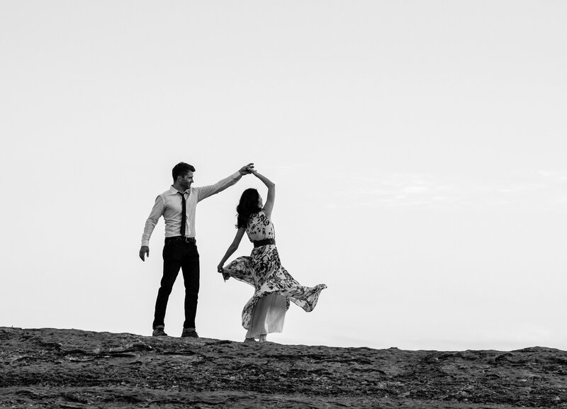 Black and white photo of couple dancing, with him twirling her in a joyful moment during their engagement session at Pedernales Falls State Park.