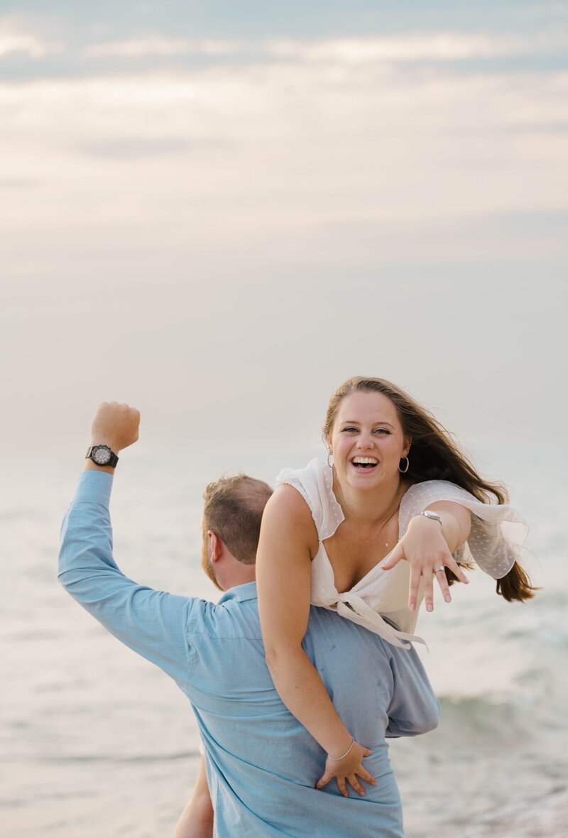 a bride shows her engagement photographer her diamond ring by Lake Michigan