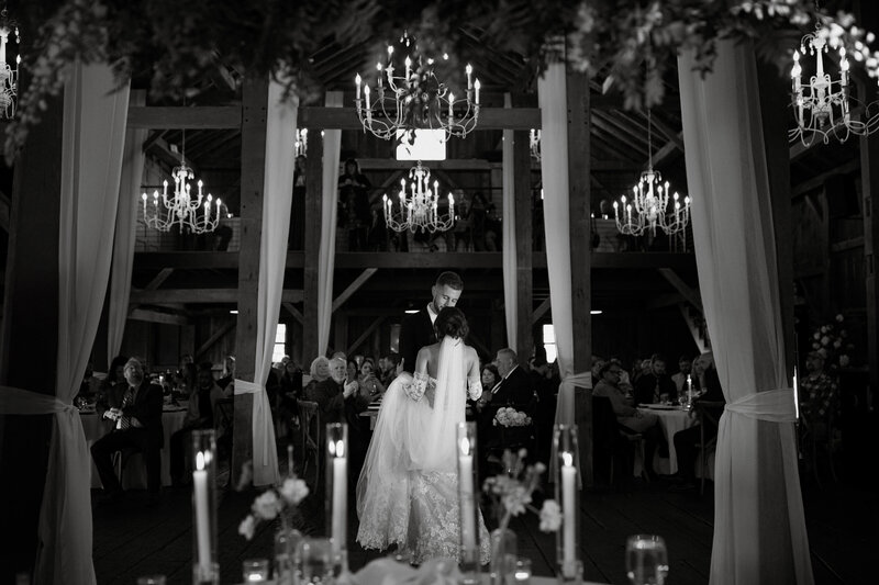 Black and white photo of a bride and groom sharing their first dance under chandeliers at a barn wedding reception in Michigan City, captured by Rainstorm Photo & Video in an emotive, cinematic style.