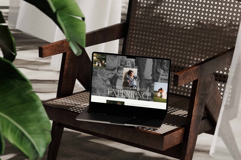 A laptop displaying a photography website designed by a skilled photography website designer is placed on a wooden chair with a cane backrest. A large green plant is partially visible in the foreground.